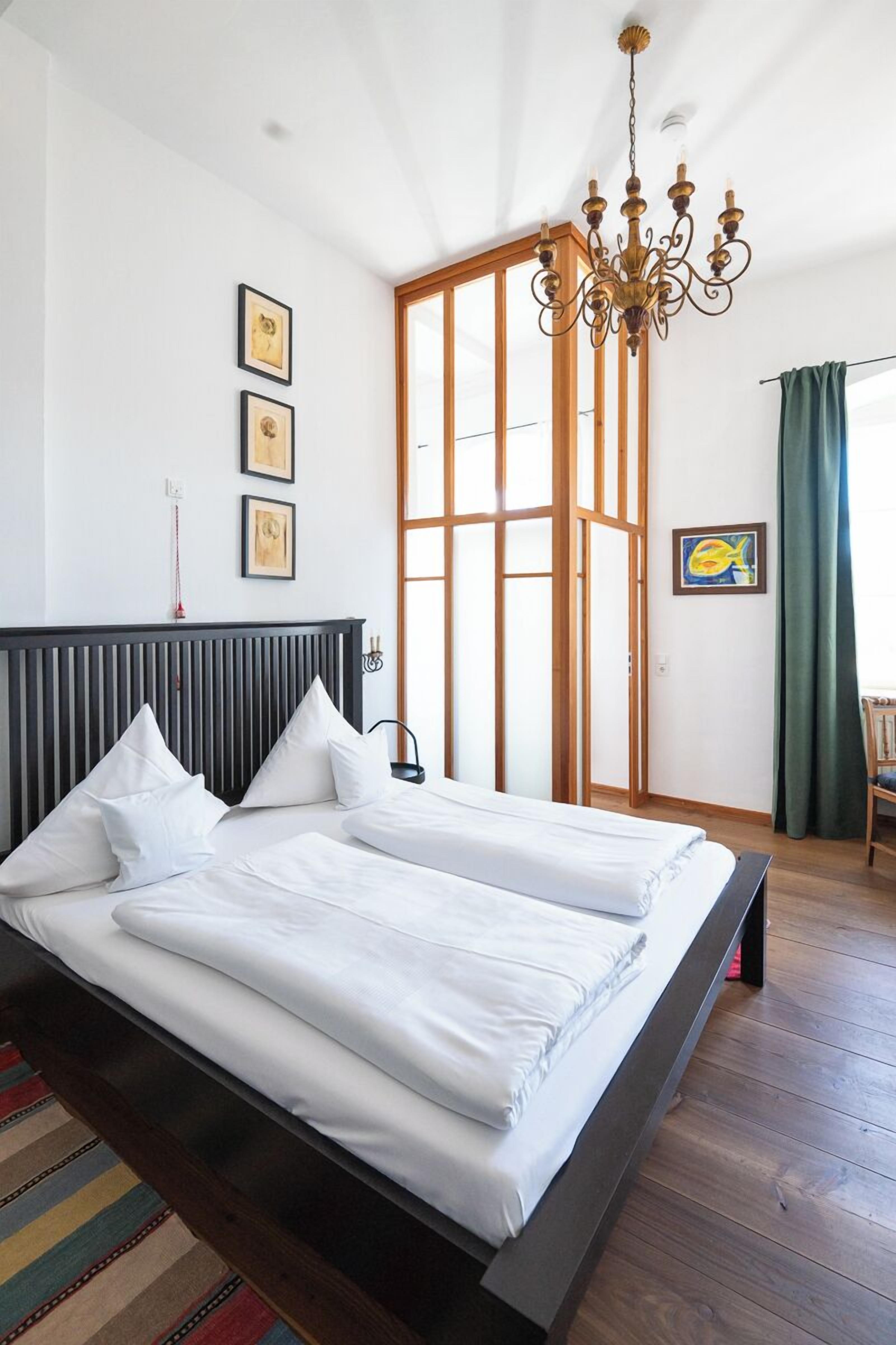Une chambre avec un grand lit, un plancher en bois, un lustre et une porte coulissante donnant sur un balcon.