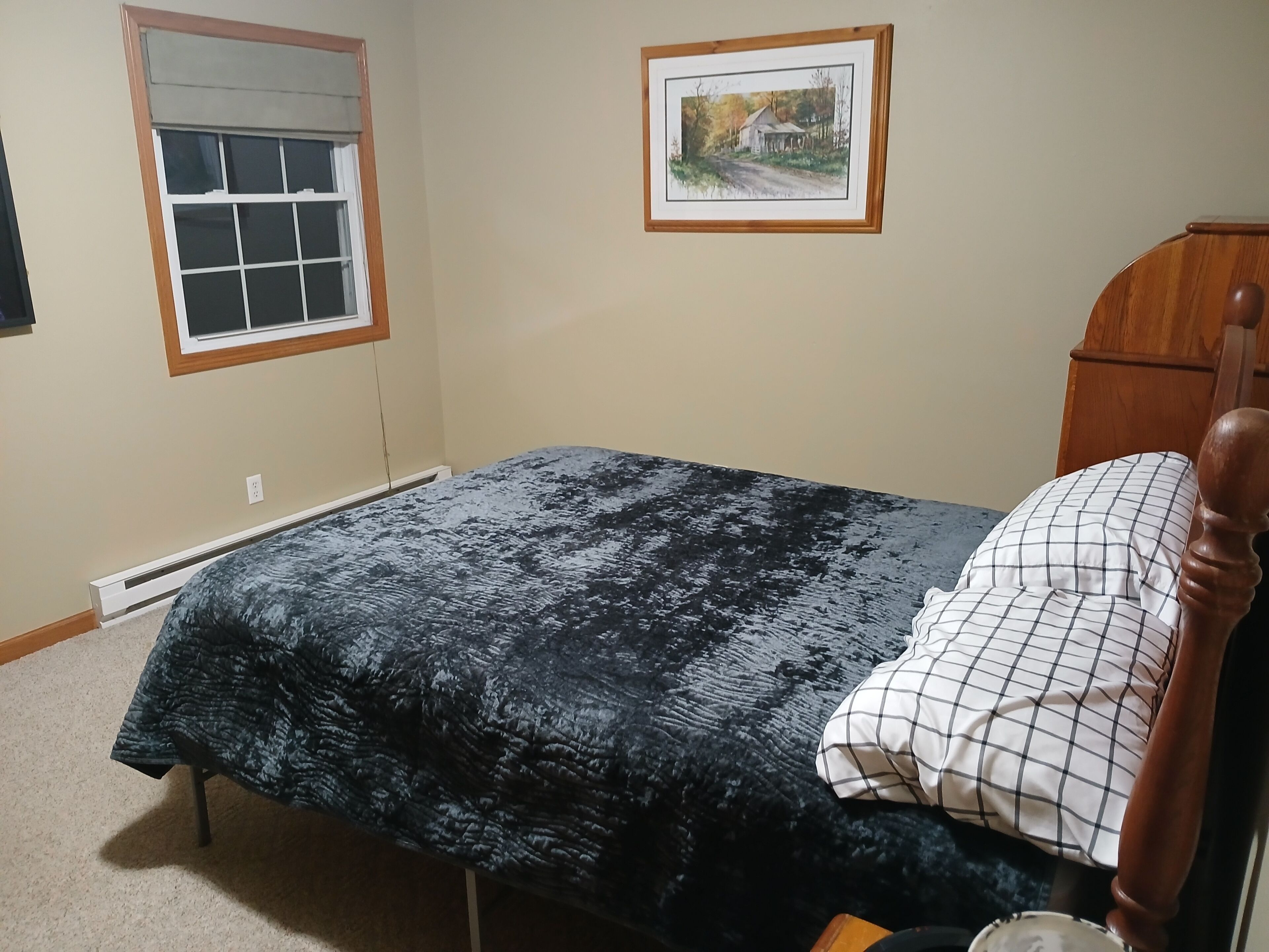 A bedroom with a bed, a window, a radiator, a framed picture on the wall, and a wooden headboard.