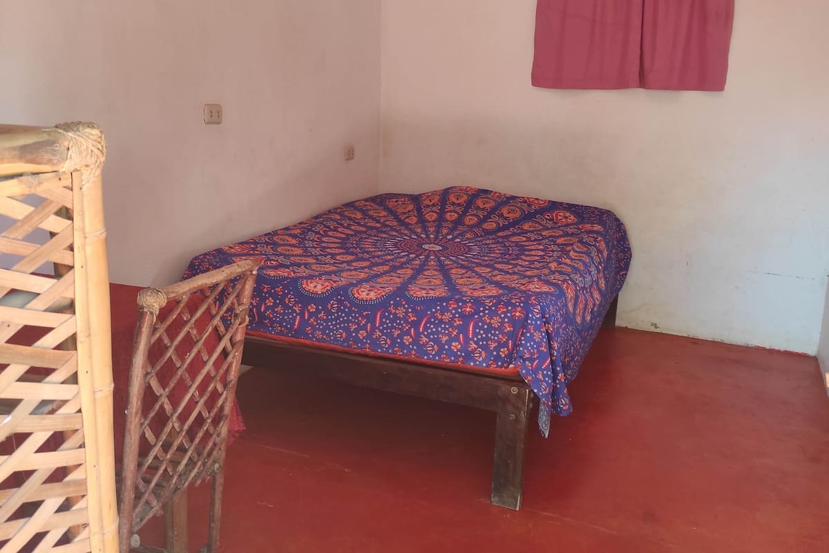 Um quarto pequeno com uma cama de solteiro, uma cadeira de madeira, piso vermelho e cortina rosa.