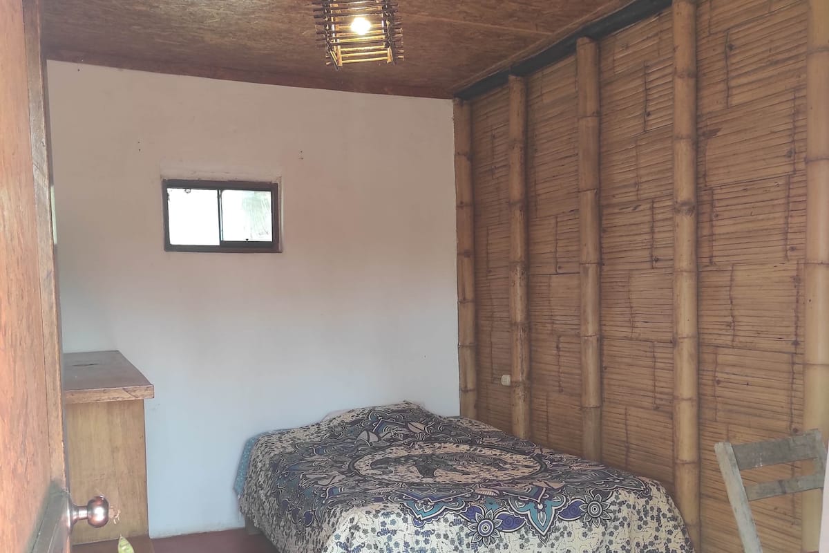 Quarto com cama, pequena janela, porta de madeira e parede de bambu.