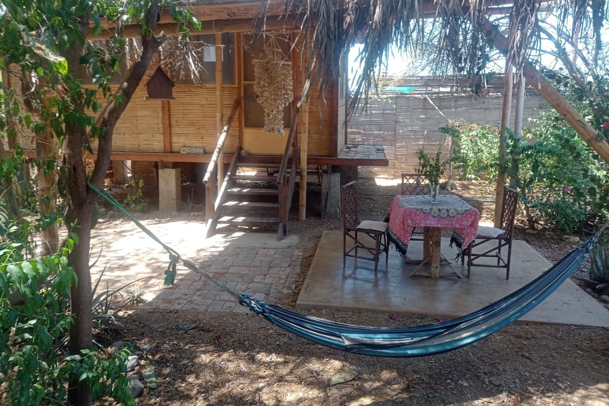 Uma cabana rústica de madeira com telhado de colmo, uma rede e uma pequena mesa com cadeiras.