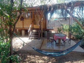 A rustic wooden cabin with a thatched roof, a hammock, and a small table with chairs. - Wasi Center (Máncora)