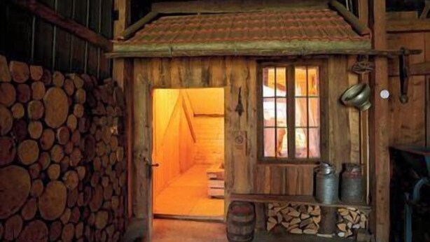 Une cabane en bois avec un toit de chaume, un escalier en bois et une petite terrasse agrémentée de barils et d’un conteneur en métal.