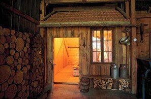Eine Holzhütte mit einem Reetdach, einer Holztreppe und einer kleinen Veranda mit Fässern und einem Metallbehälter.