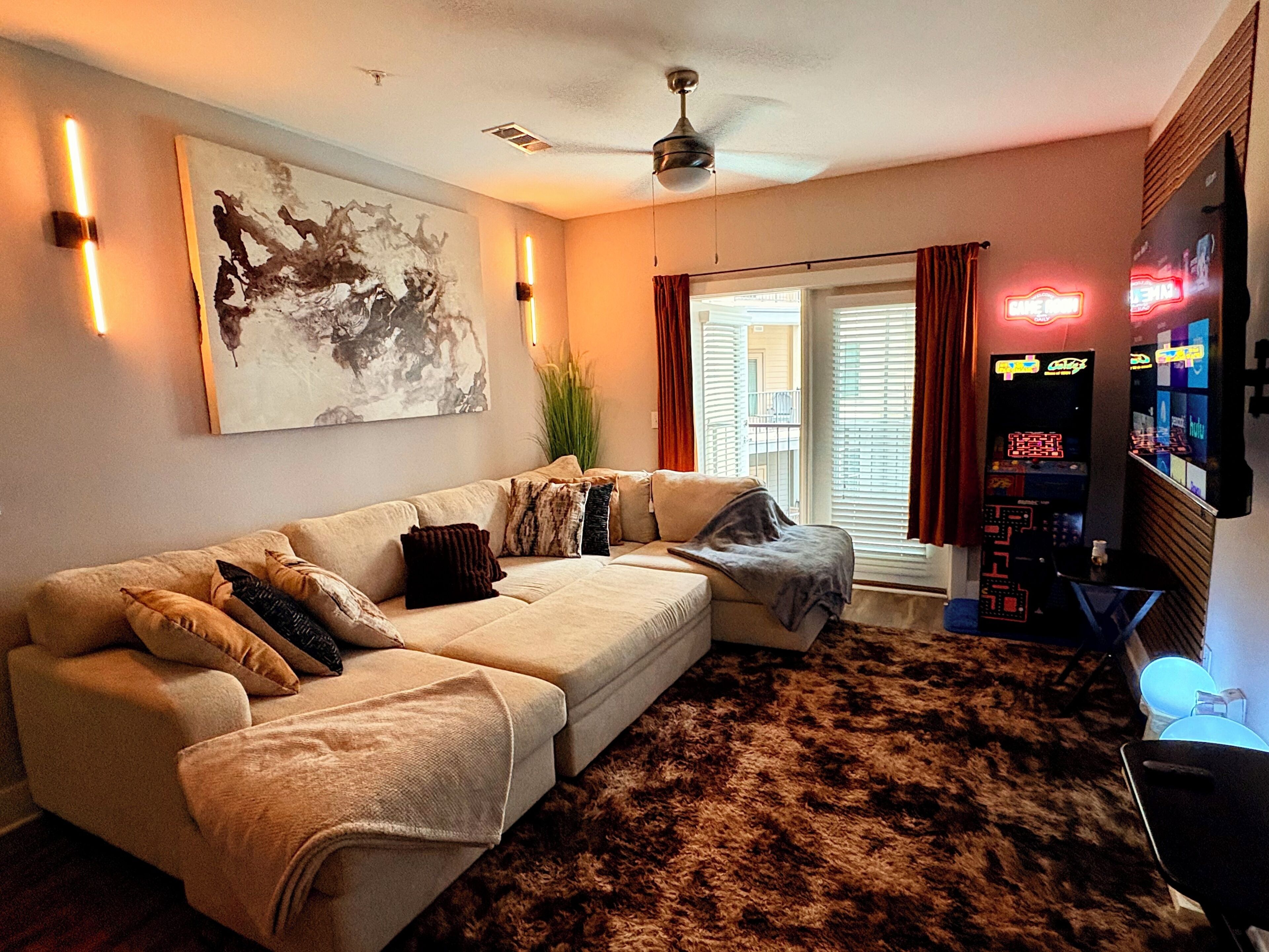 Een woonkamer met een hoekbank, een plafondventilator, een groot abstract schilderij en een bar voorzien van neonverlichting.