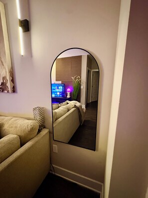 A mirror reflecting a room with a sofa, a lamp, and a wall-mounted TV.