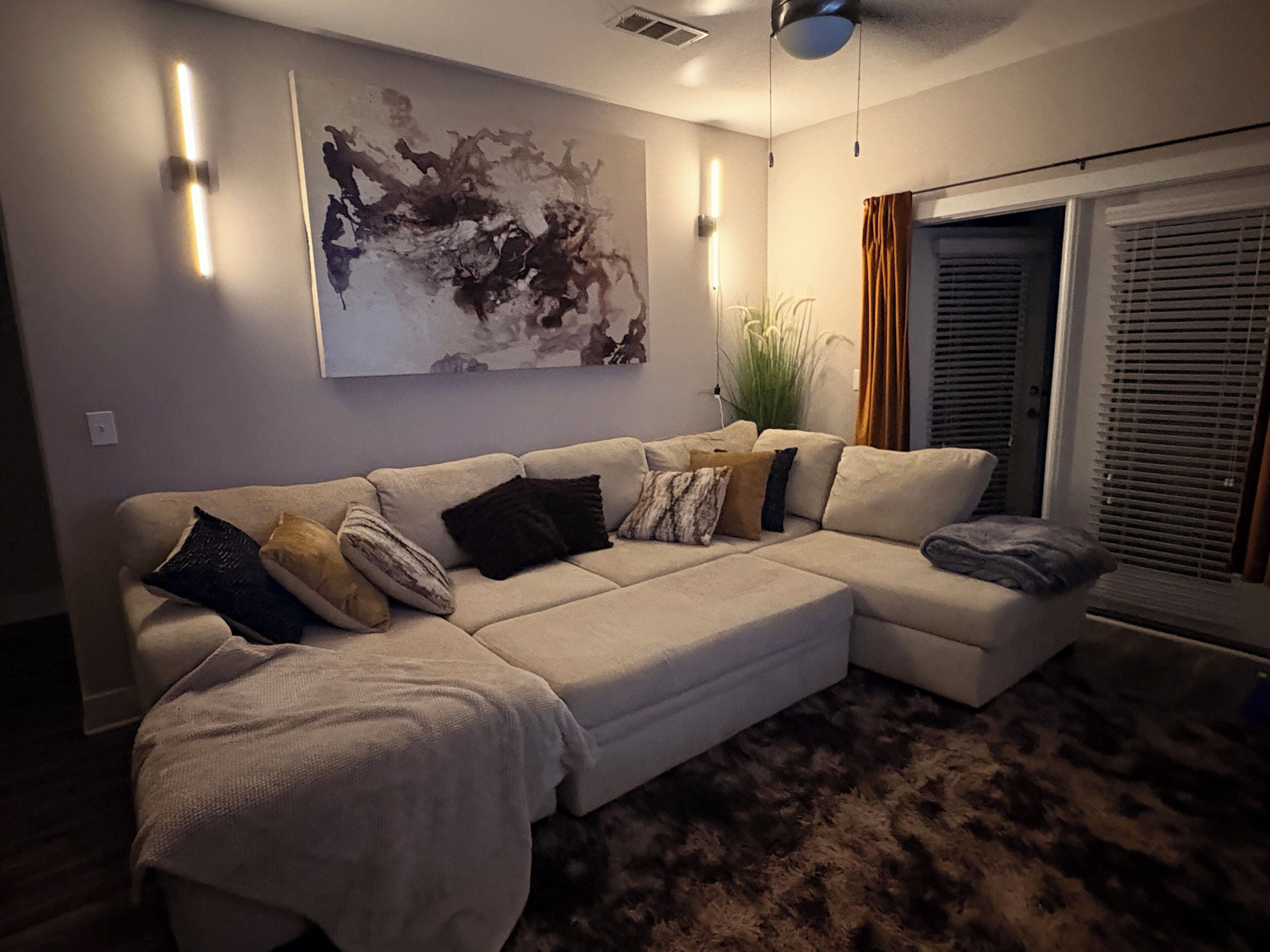 A living room with a sectional sofa, a large abstract painting, and a window with blinds.