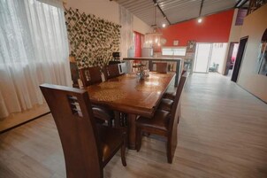 Un comedor amplio con una larga mesa de madera y múltiples sillas, un muro decorativo y una cocina al fondo.