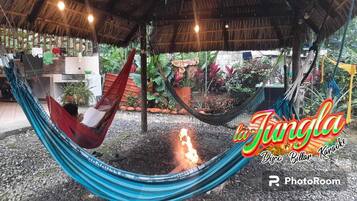 A hammock under a thatched roof, surrounded by tropical plants and a fire pit.