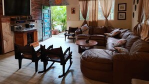 Una sala de estar acogedora con un sofá de cuero marrón, una mesa de centro de madera y un televisor montado en una pared de ladrillo. Hay dos mecedoras y una mesita con un objeto decorativo.