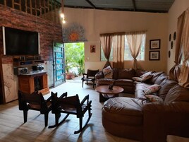 Una sala de estar acogedora con un sofá de cuero marrón, una mesa de centro de madera y un televisor montado en una pared de ladrillo. Hay dos mecedoras y una mesita con un objeto decorativo.