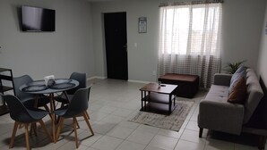 Un salon comprenant une table à manger, des chaises, un canapé, un téléviseur et une fenêtre avec des rideaux.