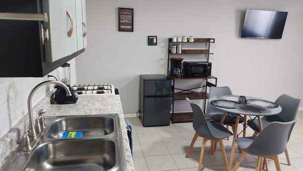 Une cuisine compacte dotée d’un comptoir en granit, d’un évier en acier inoxydable et d’un petit réfrigérateur. On y trouve également un coin repas avec une table ronde et quatre chaises. Un téléviseur à écran plat est fixé au mur, au-dessus de la table à manger.