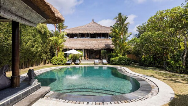 A pool with a thatched-roof structure in the background, surrounded by lush greenery and a clear sky.