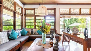 A cozy room with a wooden ceiling, large windows with thatched roofs, a sofa with blue cushions, a wooden coffee table with a vase and candles, and a statue on the table.