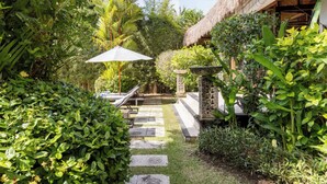 A lush garden with a thatched-roof structure, stone steps, and a pathway leading to a seating area with a white umbrella.