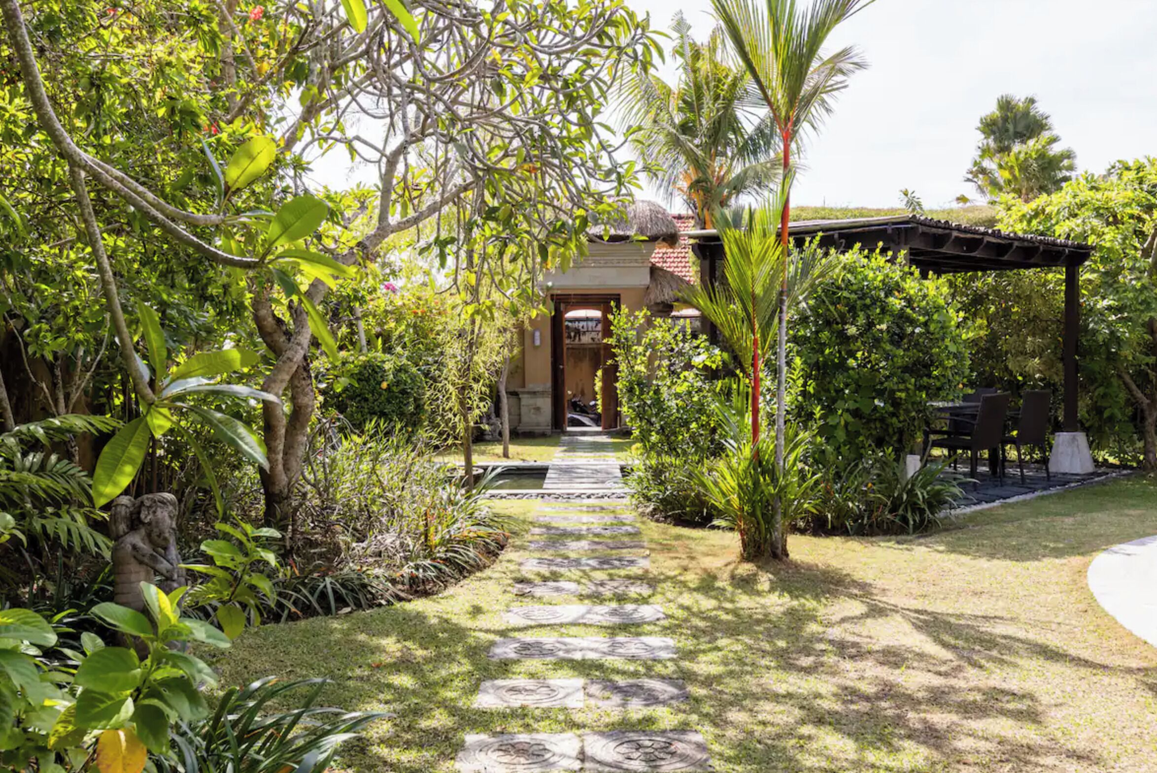 A garden pathway leading to a building with a thatched roof, surrounded by lush greenery and palm trees.