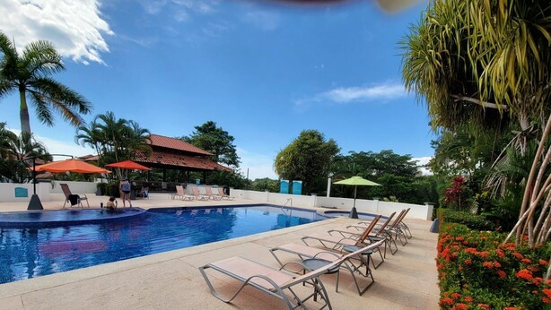 Piscina con sillas de descanso y sombrillas, rodeada de palmeras y una zona de estar cubierta.