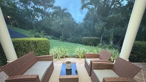 Un patio amueblado con mobiliario de mimbre y una mesa central, rodeado de vegetación y vistas a un jardín ajardinado.