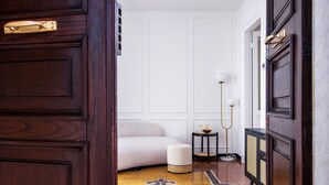 Um corredor com uma porta de madeira escura, um banco estofado branco, uma pequena mesa redonda com abajur e um tapete estampado.