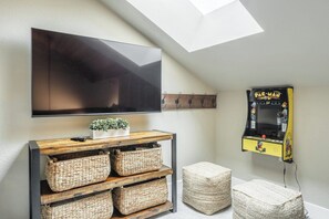 Una habitación con una televisión de pantalla plana montada en la pared, una mesita consola de madera con cestas, una máquina arcade Pac-Man y un tragaluz.