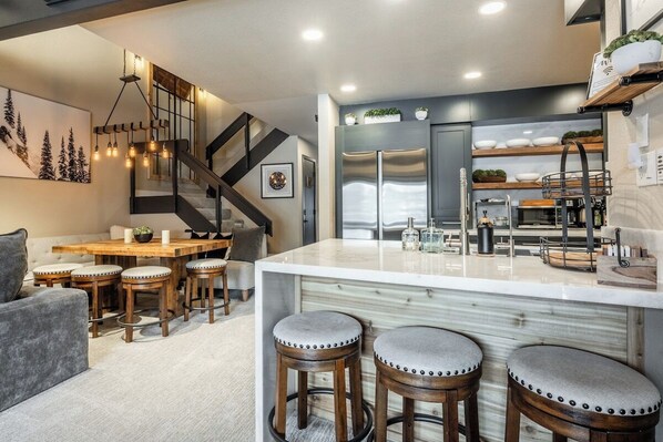 A modern kitchen with a central island, bar stools, a dining area with a wooden table, and a staircase with a dark railing.