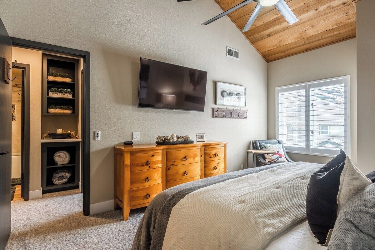 Une chambre à coucher comprenant un lit, une commode en bois, un téléviseur et une fenêtre avec des stores.