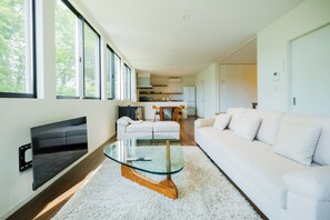 En moderne stue med et fladskærms-tv, et glasbord, hvide sofaer og et spiseområde med stole.