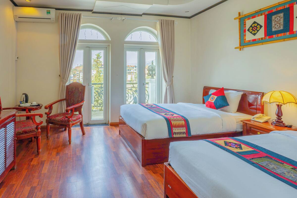 Une chambre d’hôtel avec un grand lit, un lit plus petit, un bureau avec une chaise et un balcon avec des rideaux.