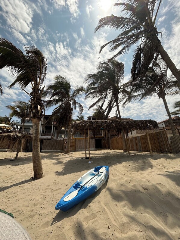 A sandy beach with a blue kayak, palm trees, and a building with a thatched roof in the background.