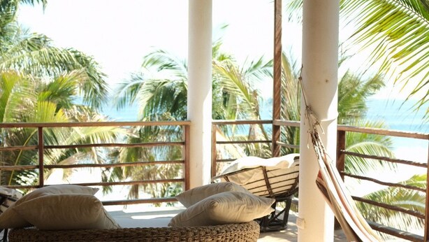 Ein überdachter Außenbereich mit Rattan-Sofa und Kissen, mit Blick auf einen Strand, an dem Palmen und das Meer zu sehen sind.