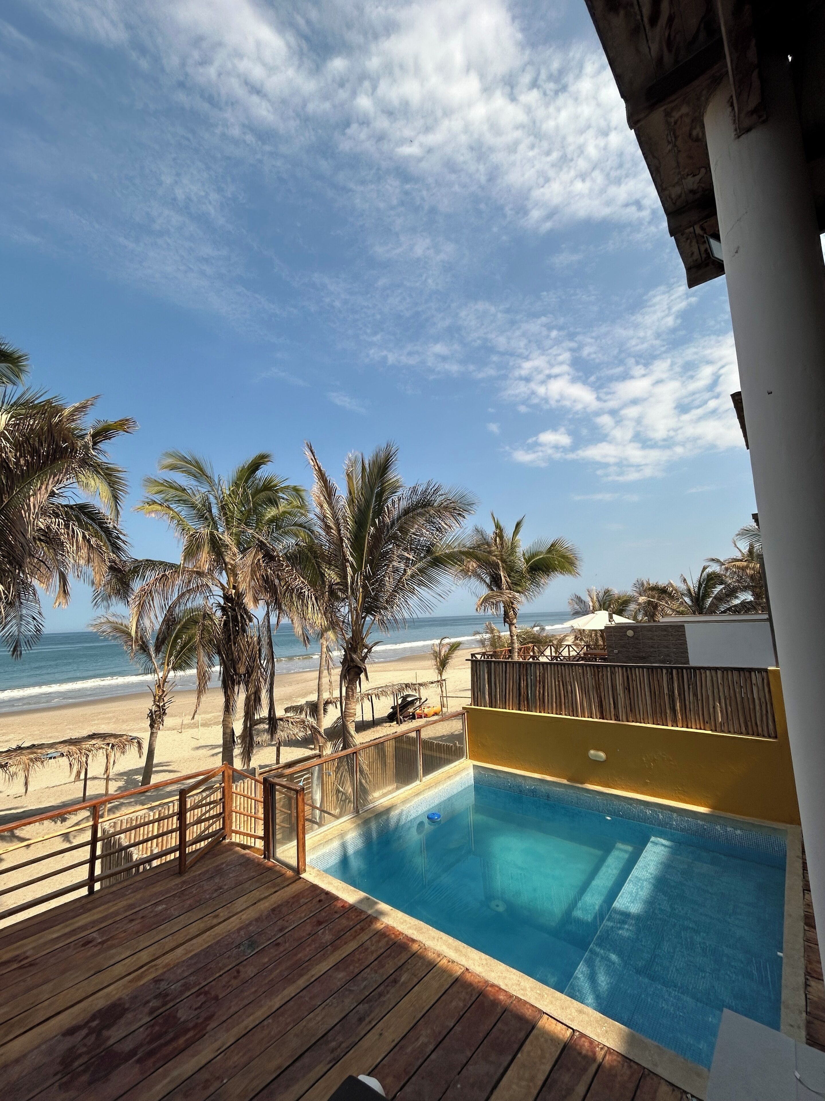 A wooden deck with a pool, overlooking a beach with palm trees and the ocean.