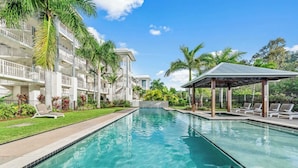 A swimming pool with lounge chairs, a covered seating area, and a multi-story building with balconies.