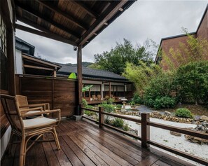 Um deck de madeira com cadeiras de vime, um caminho de pedra e vista para um edifício tradicional e vegetação exuberante.