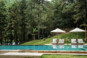 Una piscina con sillas de descanso y sombrillas, rodeada de un área boscosa.