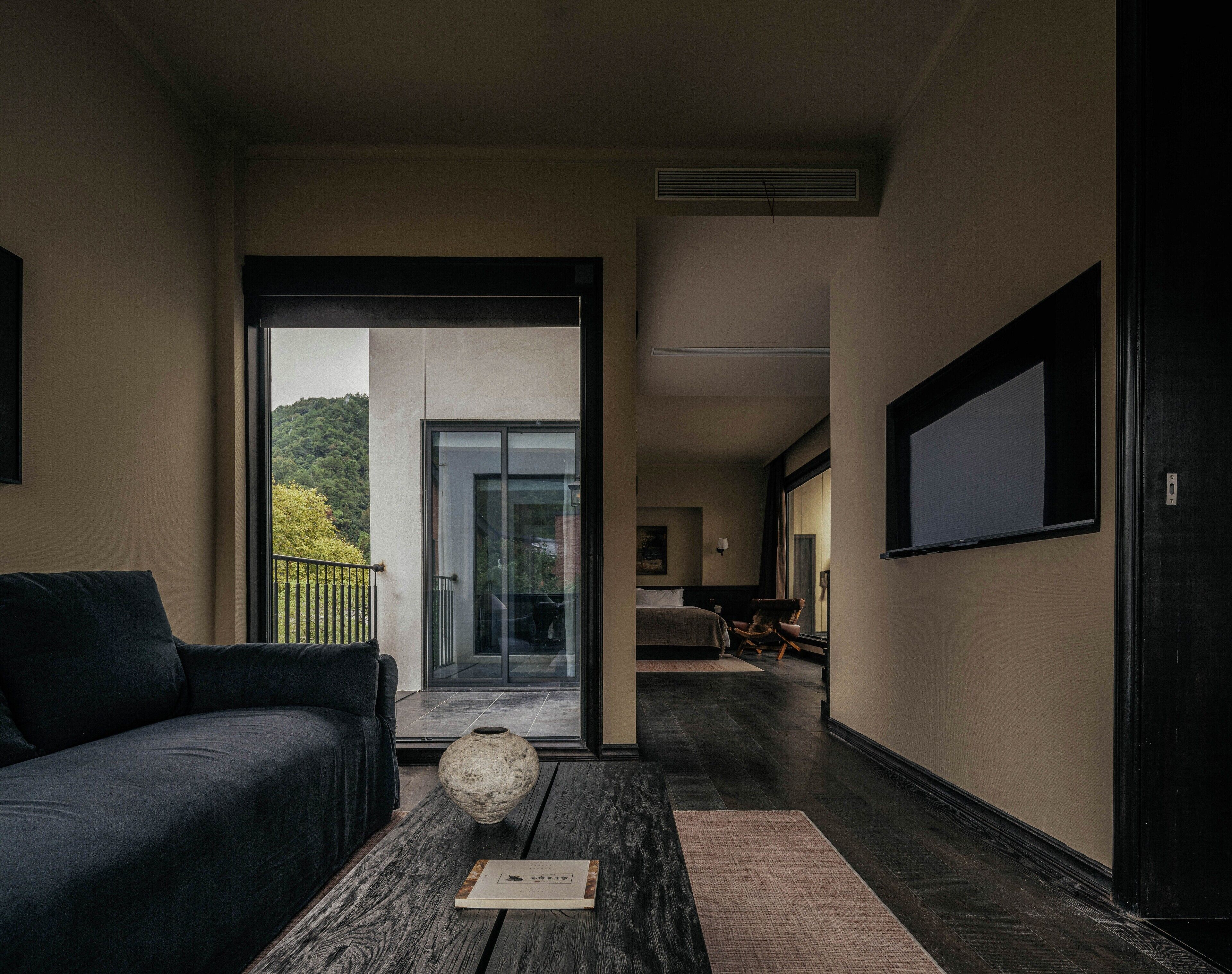 A modern hotel room with a dark-colored sofa, a wooden coffee table with a vase, and a view of a lush green landscape through a sliding glass door.