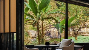 A room with tatami flooring, a low table, and a cushioned chair, with a large window offering a view of a garden with large plants and a stone wall. - Jingdezhen Xingshan City outside Jielu (Jingdezhen)