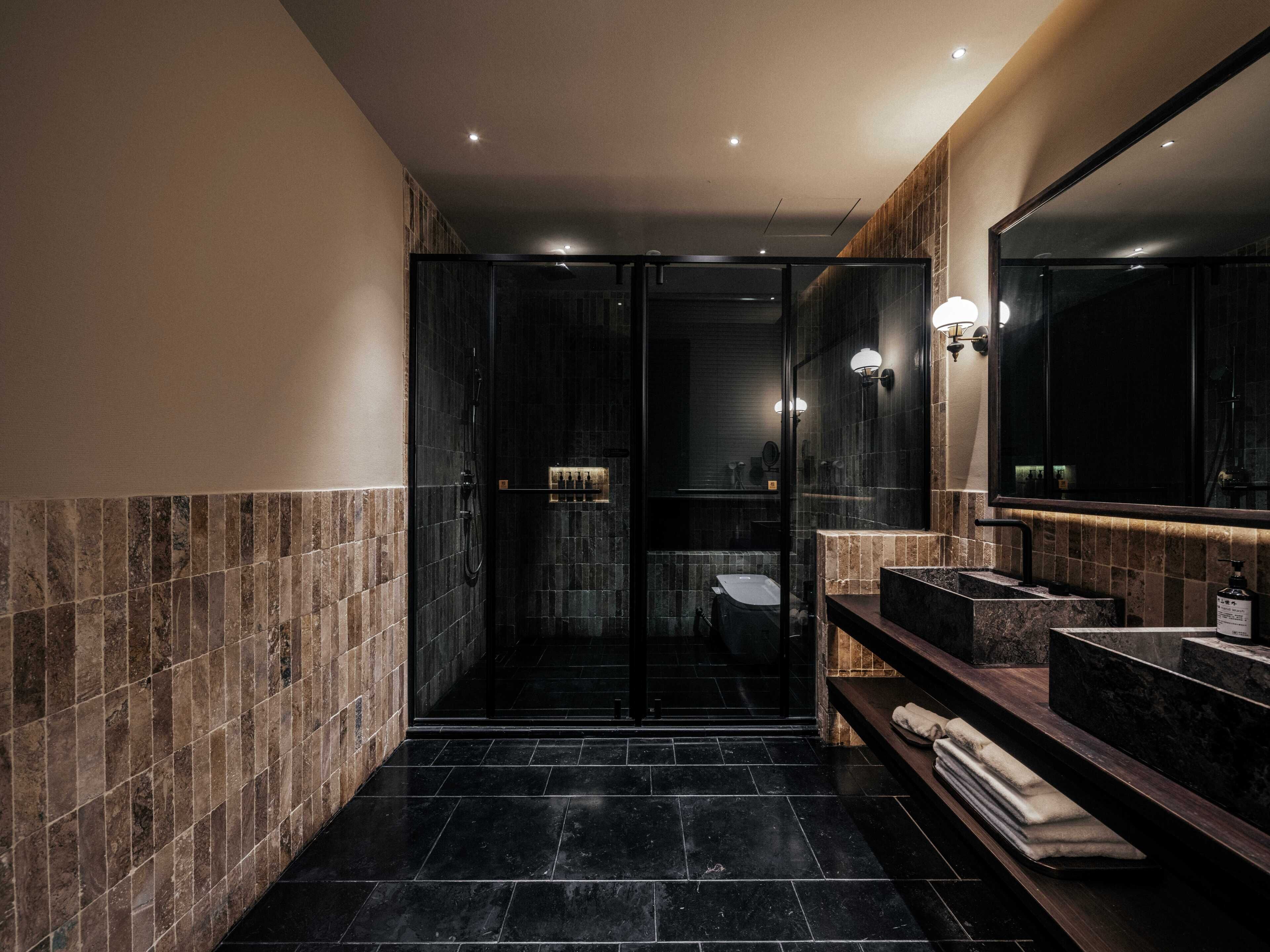 Un baño moderno con cabina de ducha de vidrio, paredes de azulejos oscuros y un espejo grande sobre el lavamanos.