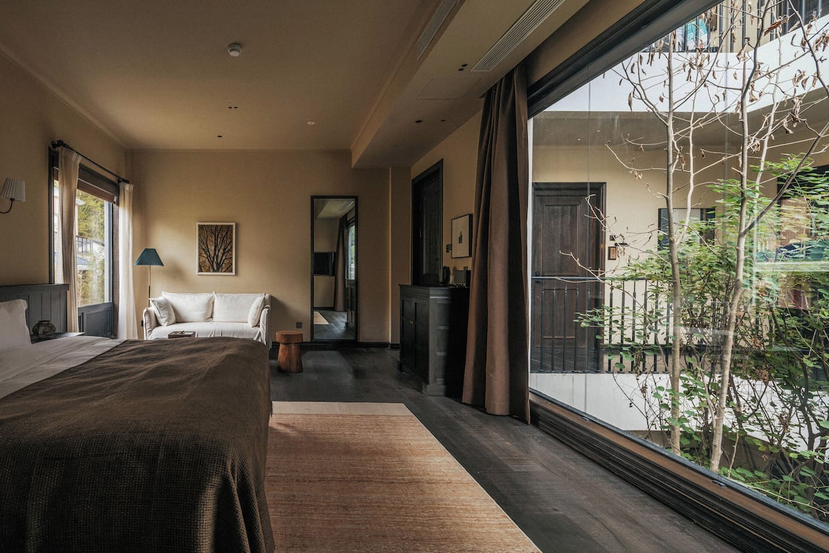 A bedroom with a large bed, a smaller bed, a nightstand, a lamp, a mirror, and a view of the outdoors.