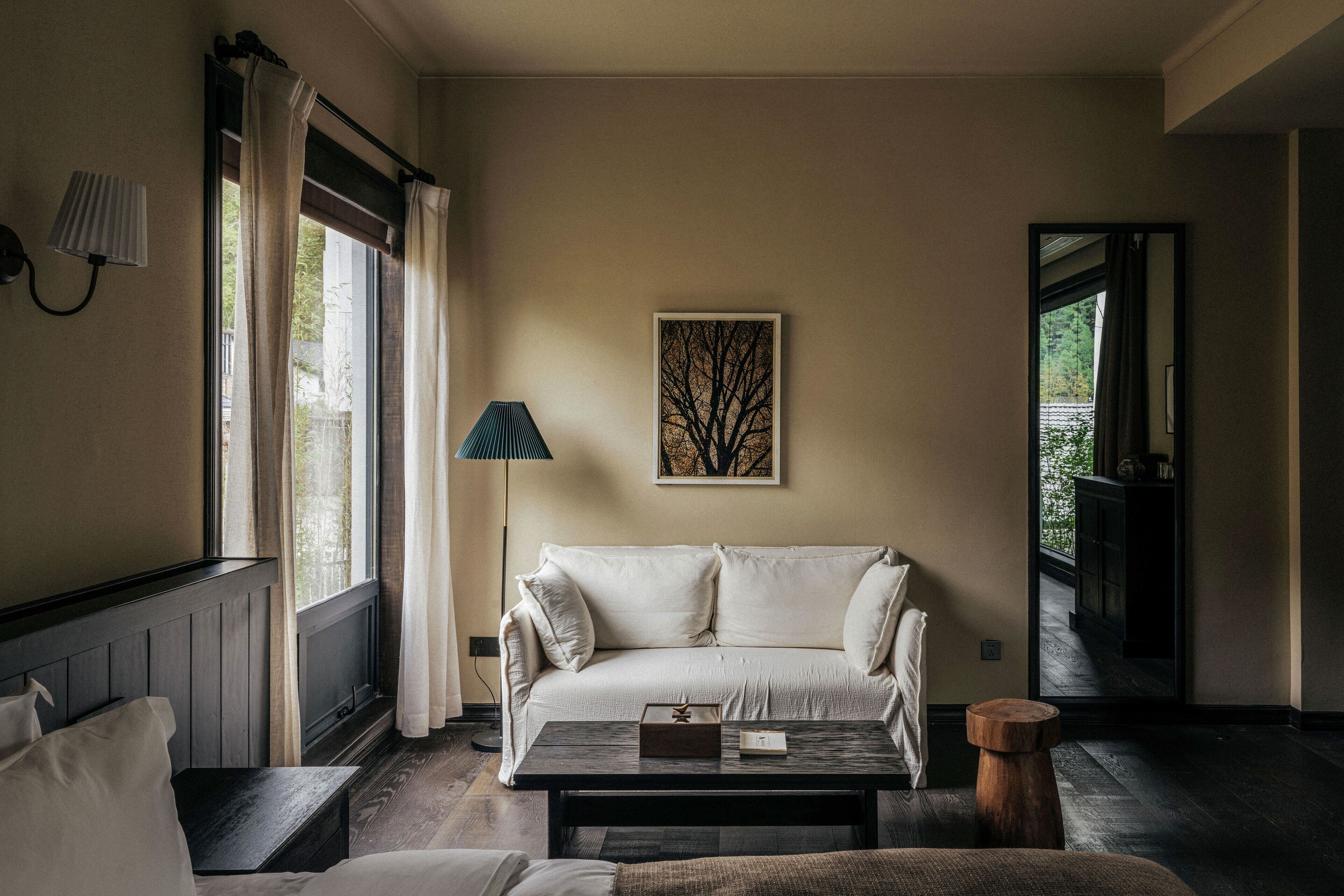 A cozy living room with a white sofa, a coffee table, and a lamp. There is a painting on the wall and a window with curtains.