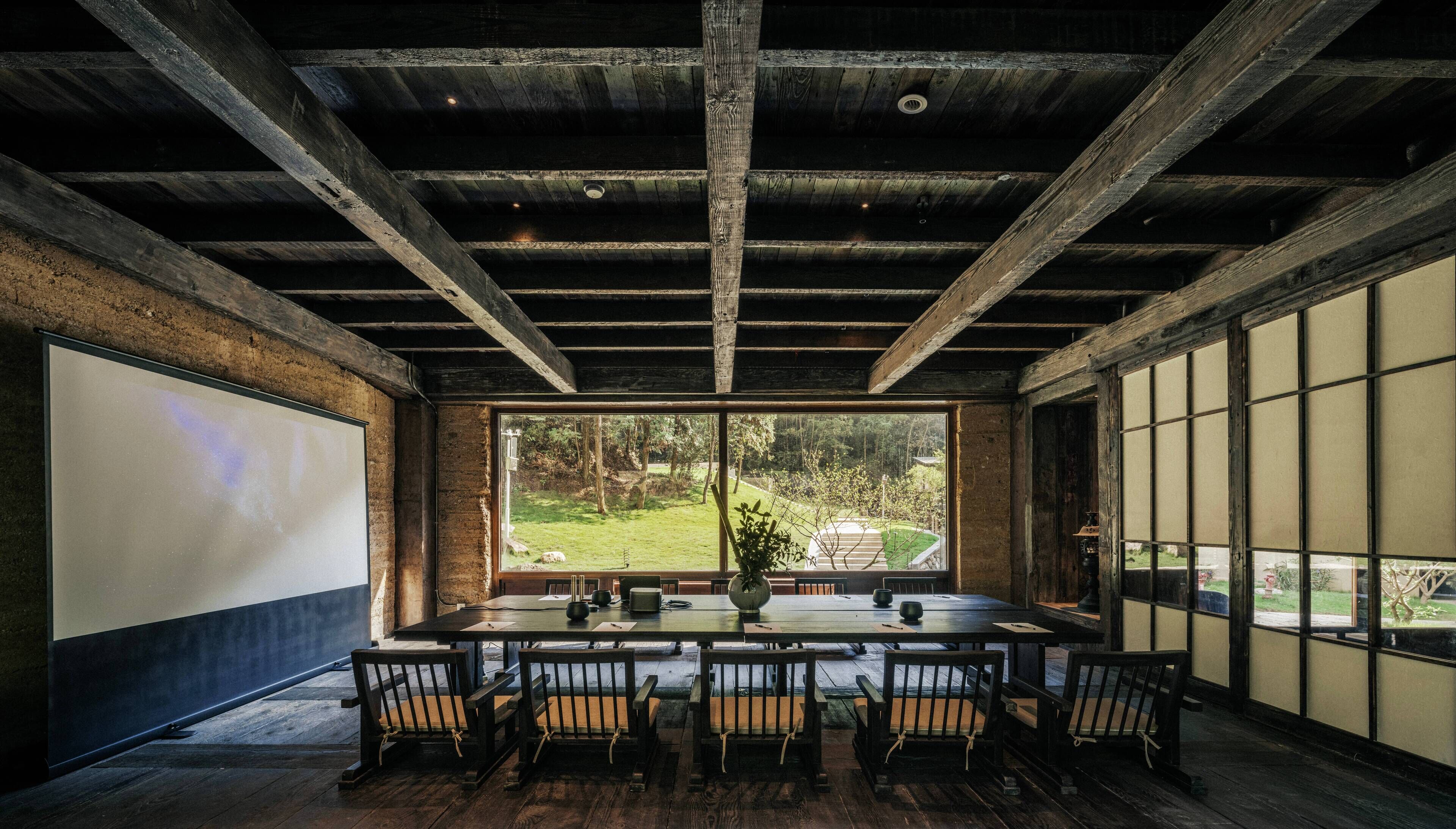 A rustic dining area with a long table and chairs, a projector screen, and large windows offering a view of the outdoors.