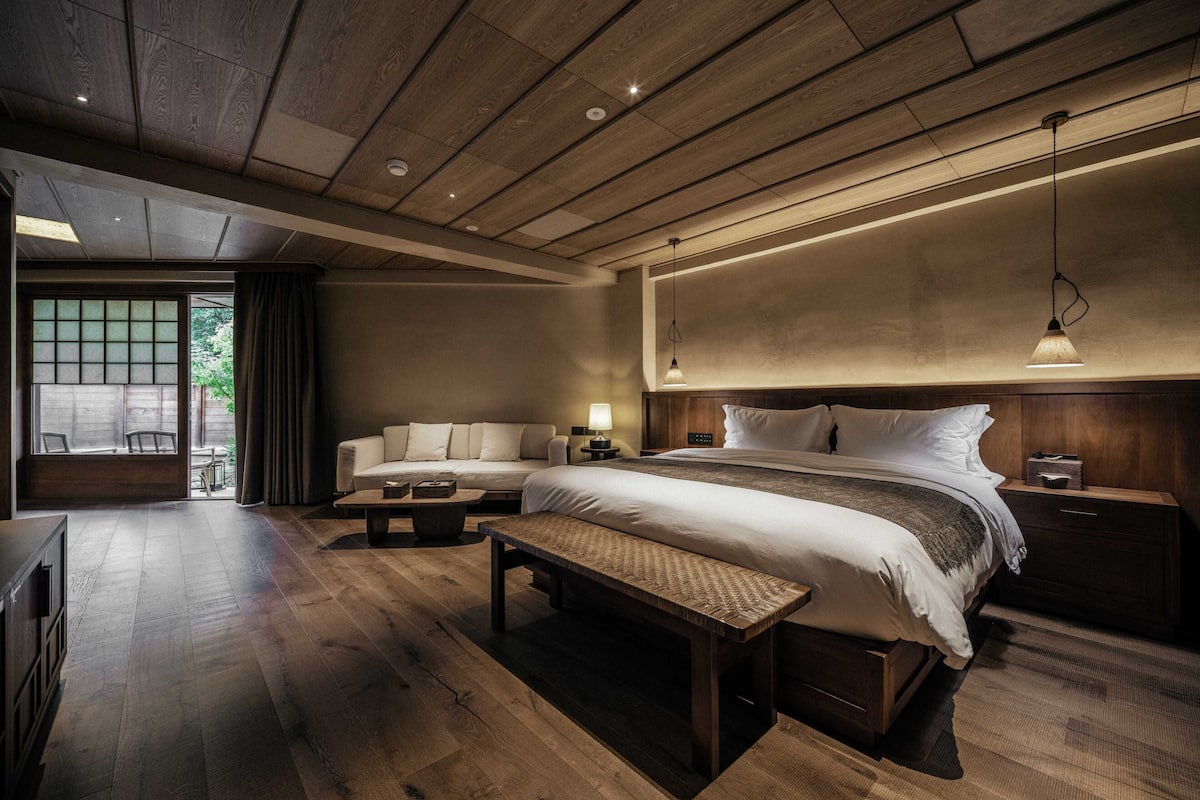 A modern hotel room with a wooden ceiling, a large bed with white bedding, a bench, and a small table.