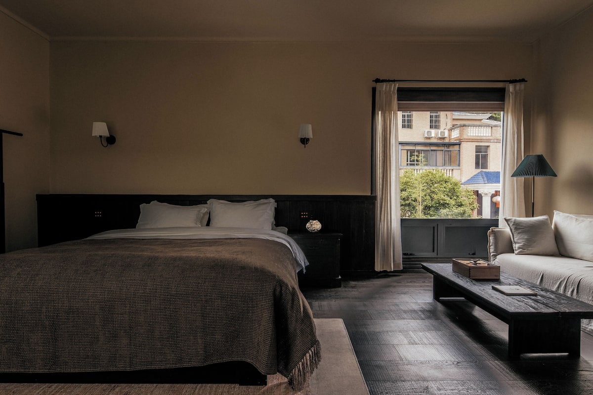 A hotel room with a large bed, a nightstand, a wooden bench, a sofa, and a view of buildings through the window.