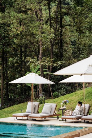 Uma área à beira da piscina com espreguiçadeiras e guarda-sóis, emoldurada por uma paisagem arborizada.