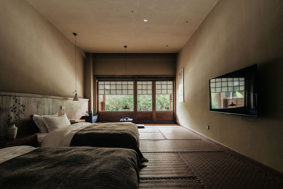 A hotel room with two beds, a television, a large window with a view of greenery, and tatami mats on the floor.