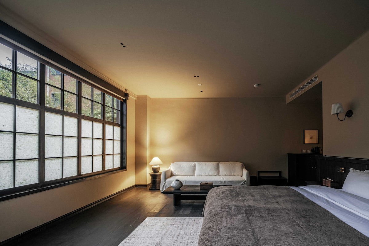 A hotel room with a large window, a bed, a sofa, a nightstand, and a lamp.