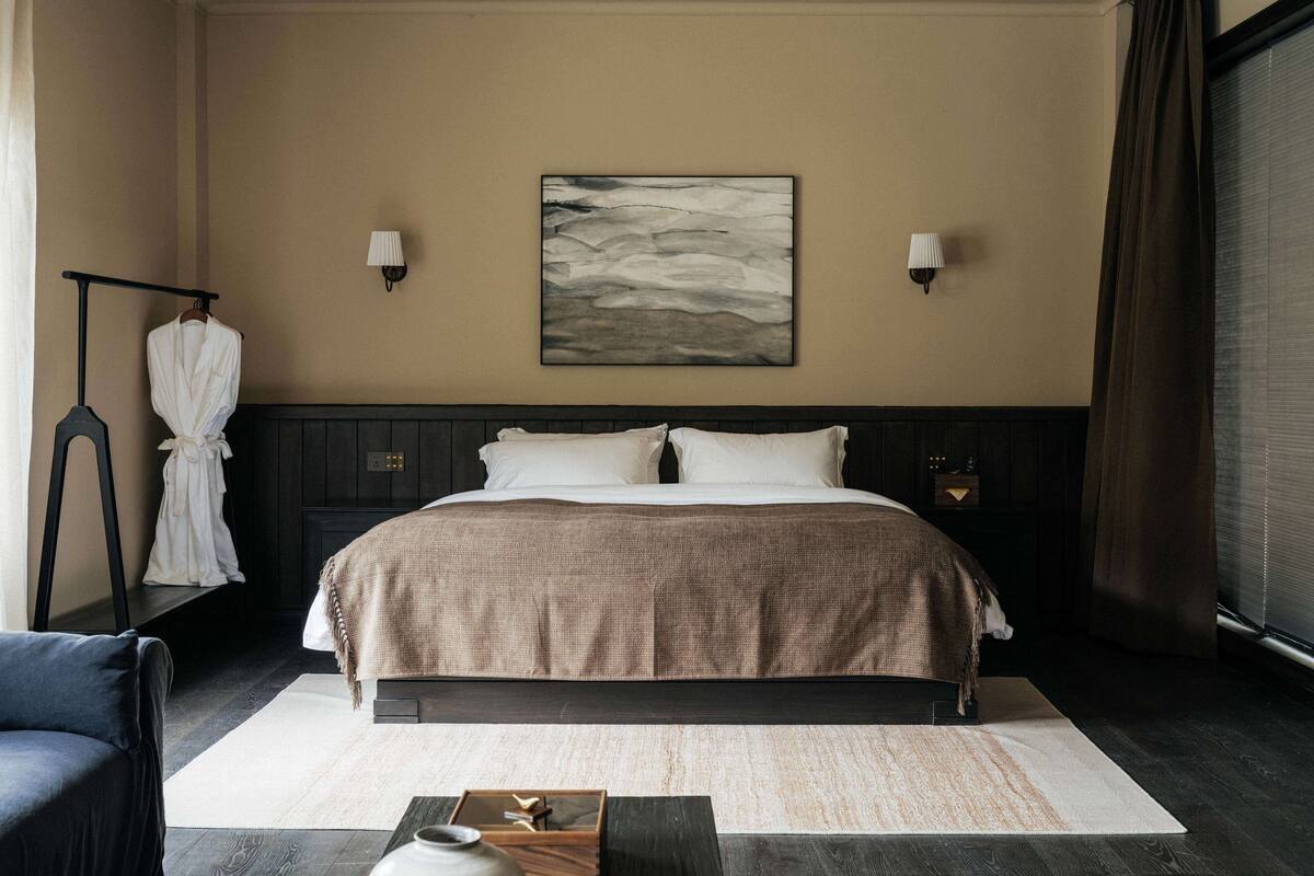 A hotel room with a large bed, two bedside lamps, a painting on the wall, a robe rack, and a desk with a vase.