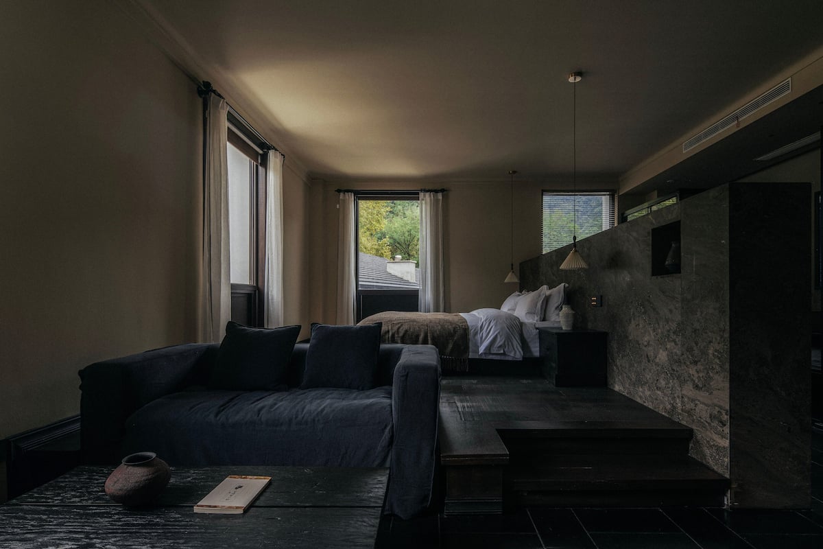 A modern hotel room with a dark-colored sofa, a small table with a vase and a book, a bed with a white headboard, and a view of trees through the window.