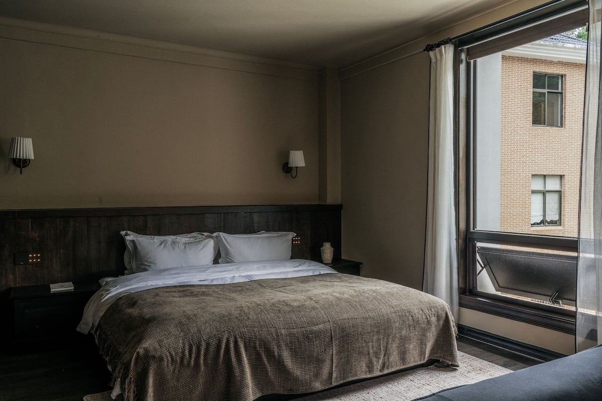 A hotel room with a large bed, a nightstand, a lamp, and a view of a building through a window.
