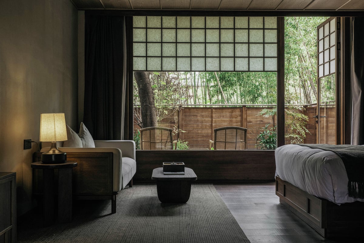A hotel room with a bed, a sofa, a small table, a lamp, and a view of bamboo outside.